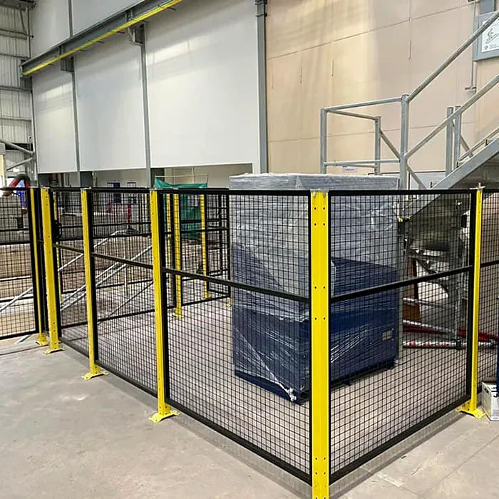 Black mesh fencing with yellow posts around a wrapped load beside industrial stairs.