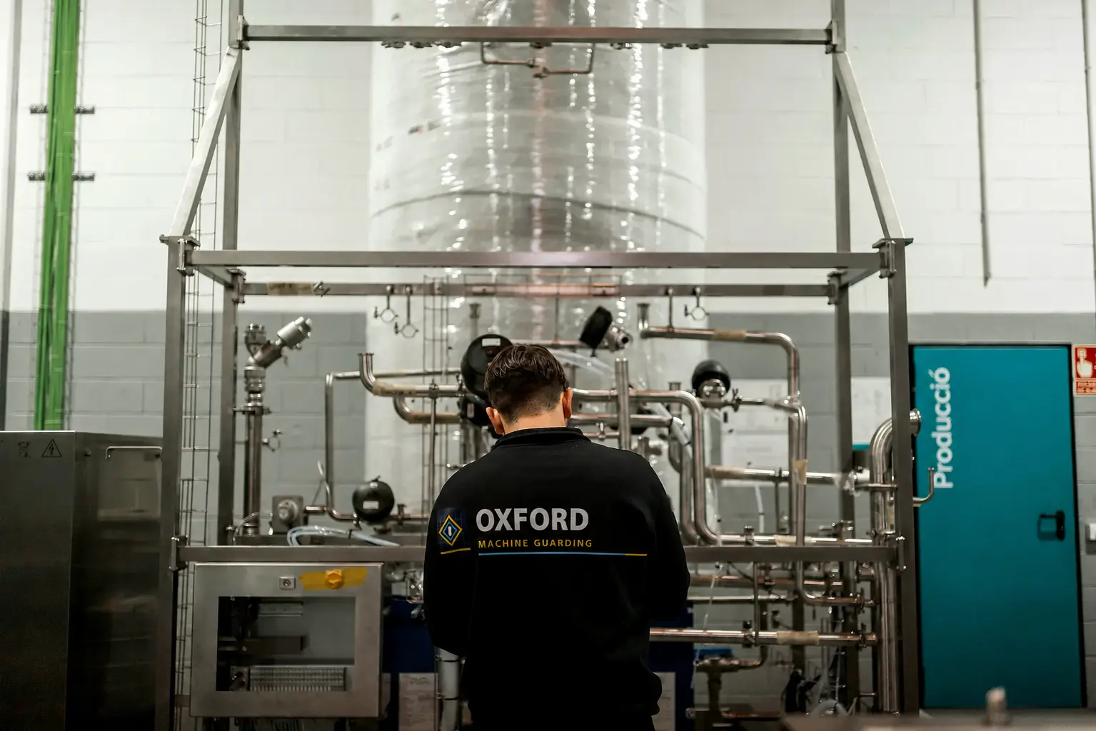 Person in a branded black shirt facing industrial pipework and equipment.